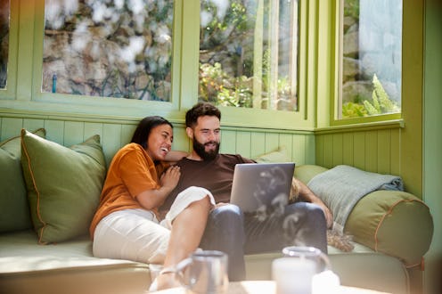 Young couple sitting on a living room sofa at home with their dog and laughing at something streamin...