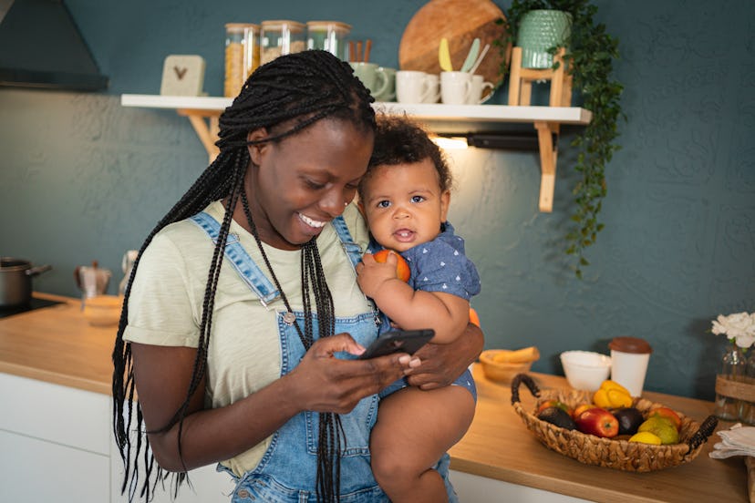 Mother Black ethnicity holding her her baby son, while using the mobile phone in the kitchen