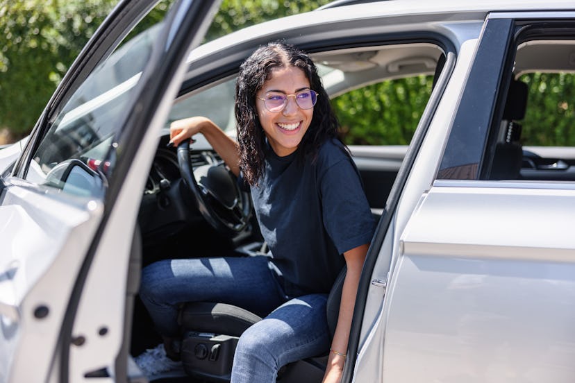 Happy young Latin female with glasses is stepping out of her parked car. The car door is open.