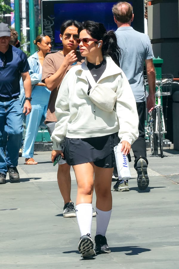 Camila Cabello wears a black pleated tennis skirt, a gray sweater, a white Lululemon belt bag, and b...