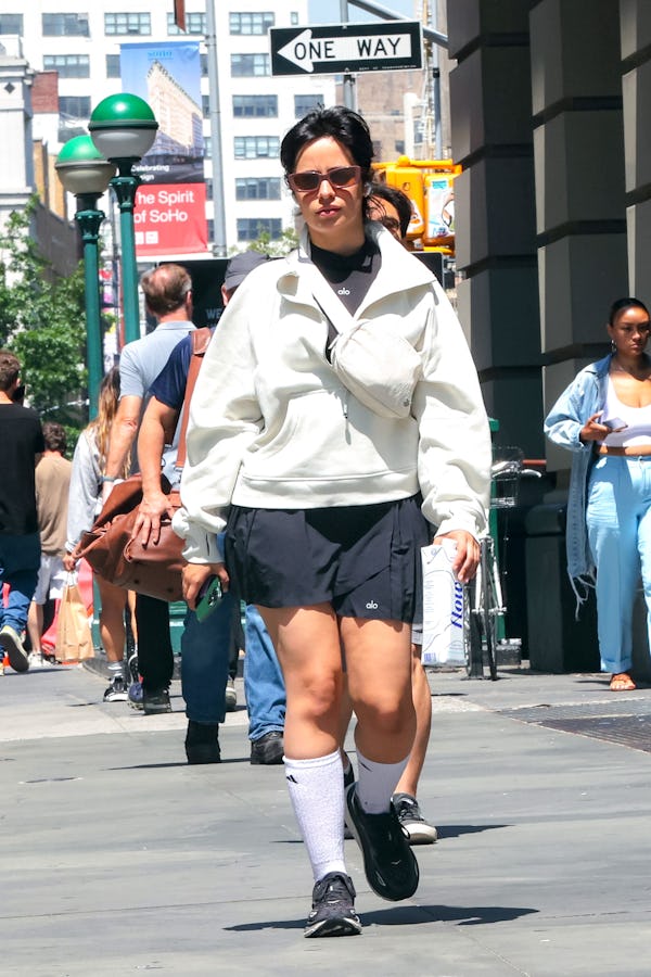 Camila Cabello wears a black pleated tennis skirt, a gray sweater, a white Lululemon belt bag, and b...