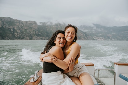 two women embrace and smile at the camera as they enjoy a boat ride, as they consider their July 202...