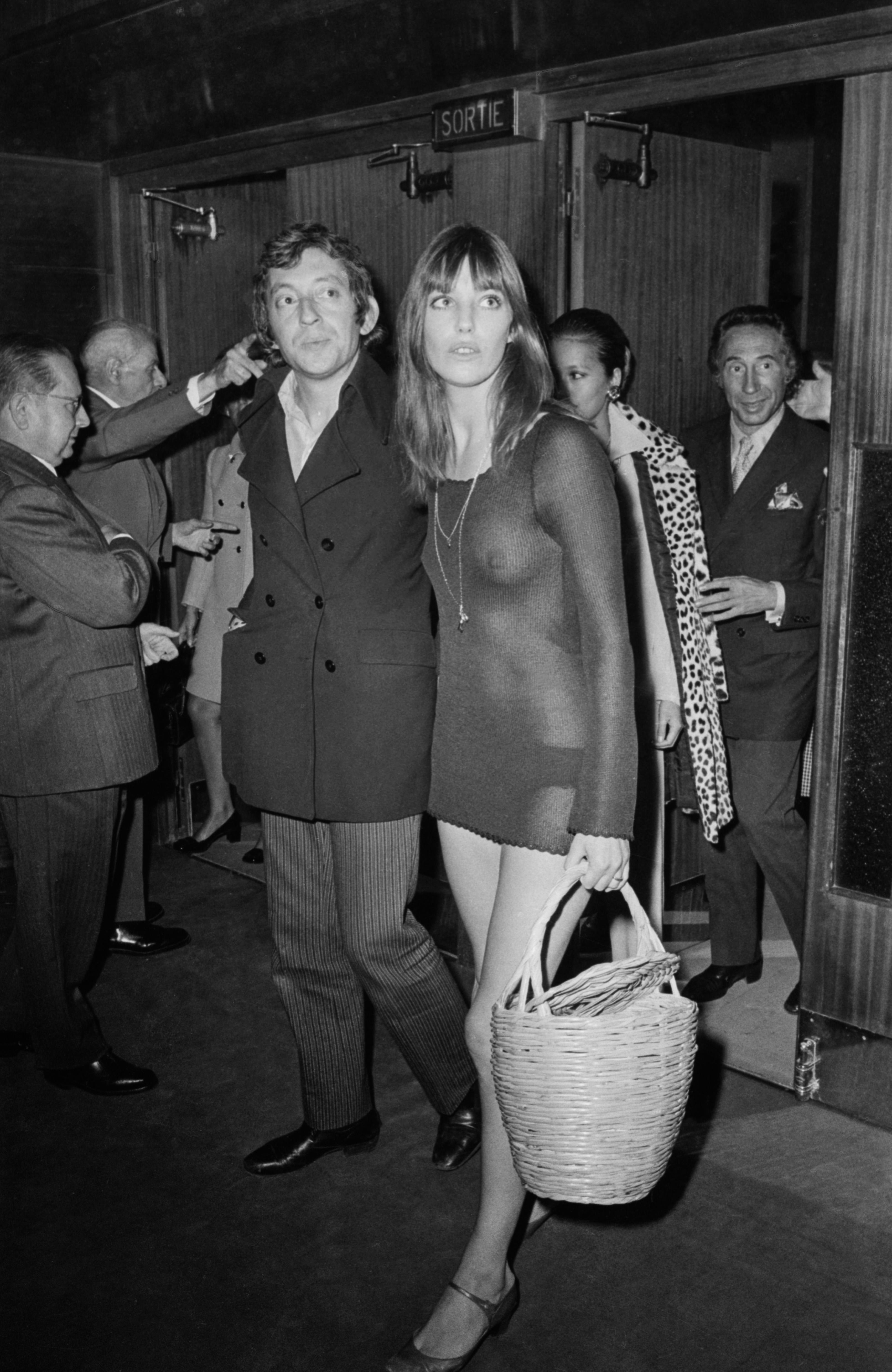 Jane Birkin wears a mini sheer dress with long sleeves, Mary Janes, and a wicker basket bag.