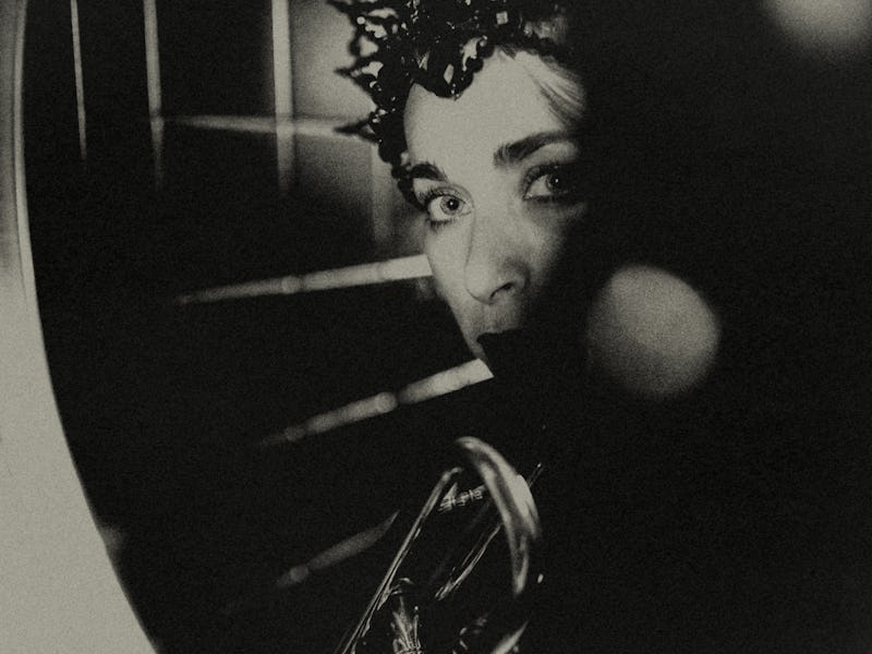 A young woman wearing a crown of pearls looks anxiously at the viewer through the window of a nightc...
