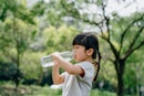 A little girl with pigtail braids drinks out of a huge clear plastic water bottle in the woods