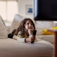 Six year old boy sitting on a couch, holding a remote control, watching tv, hand on chin