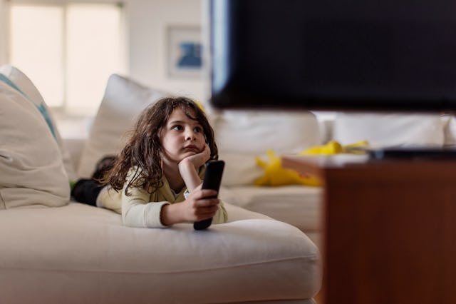 Six year old boy sitting on a couch, holding a remote control, watching tv, hand on chin