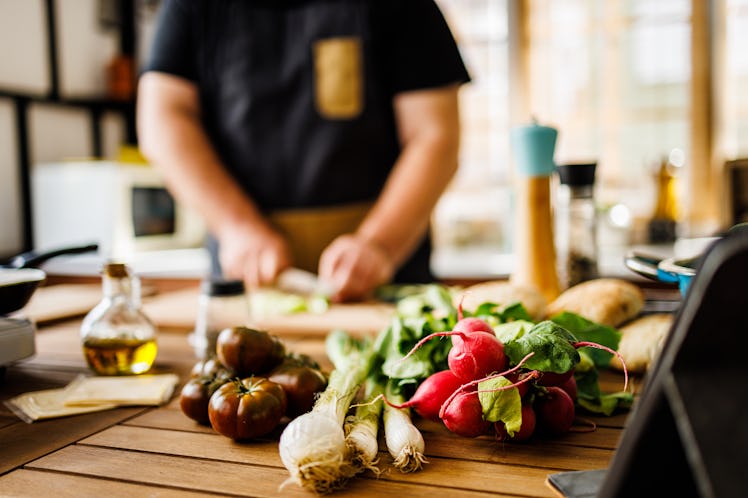 Fresh vegetables are on a kitchen counter as a man in the background chops them to prepare to eat.