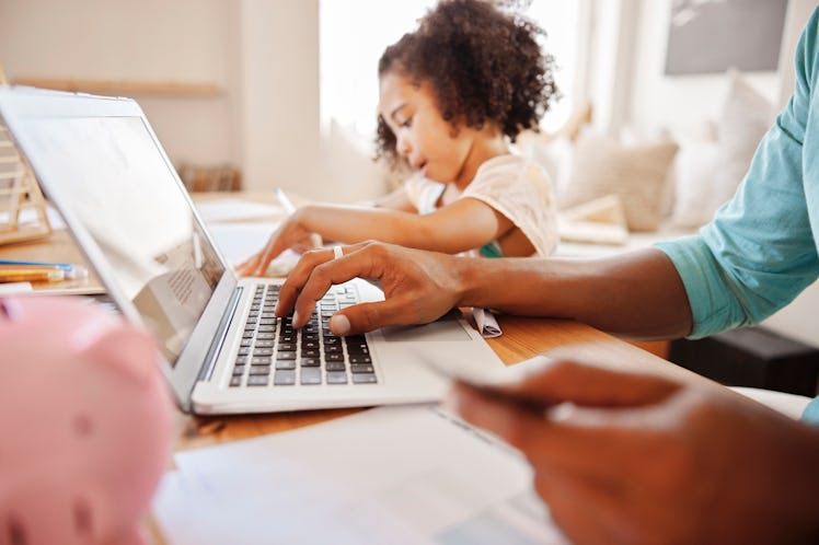 A dad on his laptop, with a piggy bank next to it and his daughter in the background.
