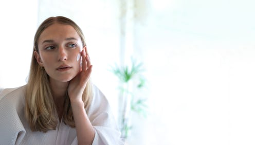 Woman gently applying face moisturiser with fingers in bathroom. Woman looking at self reflection in...