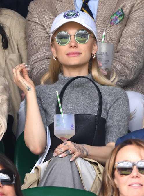 Ariana Grande wears a gray mock turtleneck top and skirt, green sunnies, and a white cap to watch th...