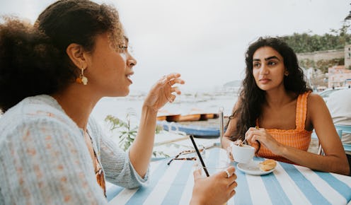 Two female friends sit at a table outdoors. They have a lighthearted discussion as they have a coffe...