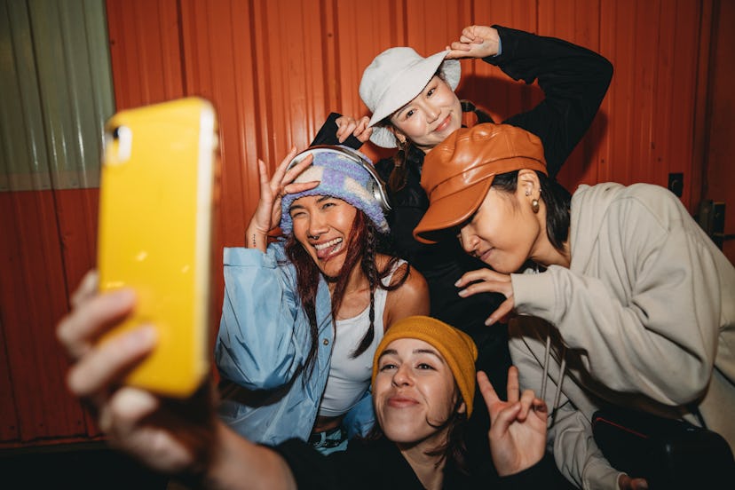 Young adult women wearing cool street style clothes are taking a selfie at night. They are wearing h...