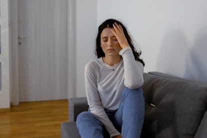 Sad depressed young woman sitting on living room sofa, touching her head and suffering from headache