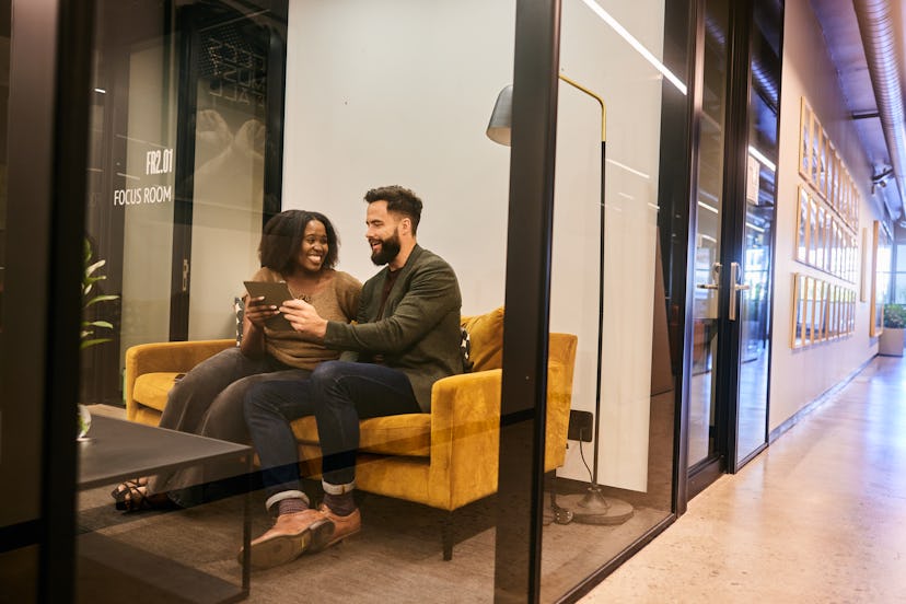 Two diverse young businesspeople laughing while working on a digital tablet on a sofa in an office m...