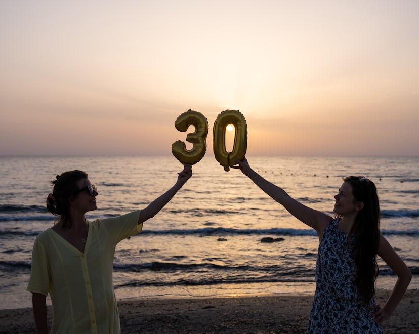 Two women celebrating birthday. Holding a large inflatable 30 sign balloon. Beach party at sunset go...