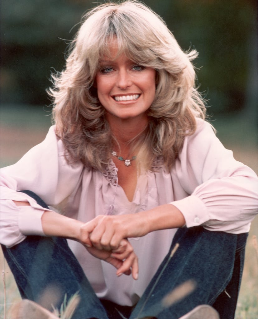 Publicity portrait of American actor and model Farrah Fawcett smiling while sitting outdoors in blue...