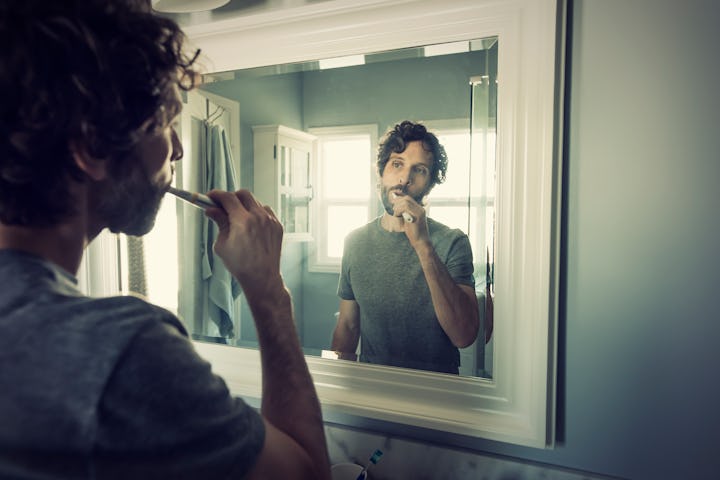 A white man with curly brown hair in a grey t-shirt brushes his teeth in the mirror. It's daylight o...