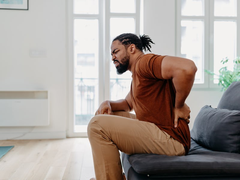 A young African American man is sitting in his living room and leaning forward from his back pain.