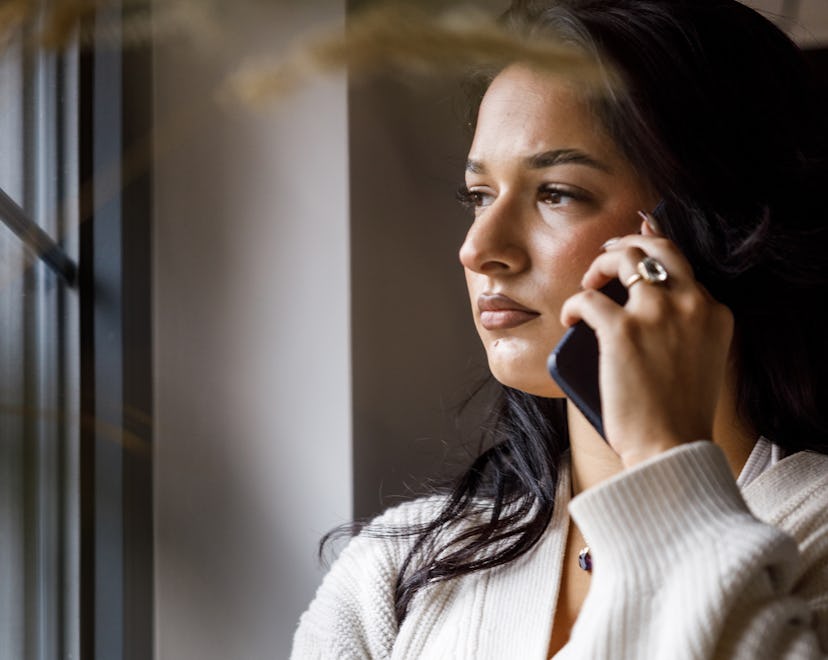Portrait of worried young woman standing by the window, arm crossed, looking outside while waiting f...