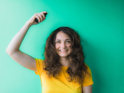 Young woman in yellow t-shirt trying to tame her frizzy curly hair and spraying hair care on it  on ...