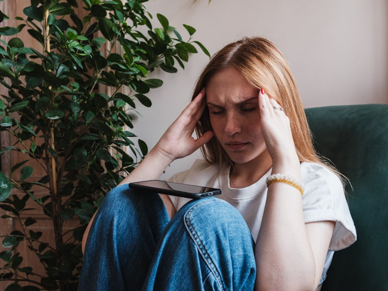 Stressed anxious young woman with smartphone, holding head in hands while sitting with closed eyes o...