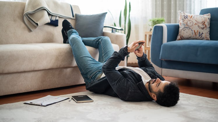 Man stretches his feet on the sofa and looks at the phone while lying on the floor