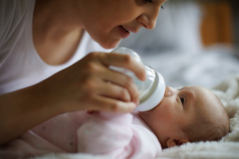 Smiling mother feeding her newborn baby girl with bottle at home, how to mix fresh and cold breast m...