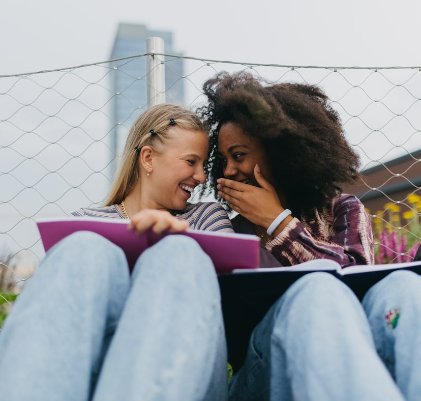 two young women laugh together as they study, and discuss why their zodiac signs will be the luckies...