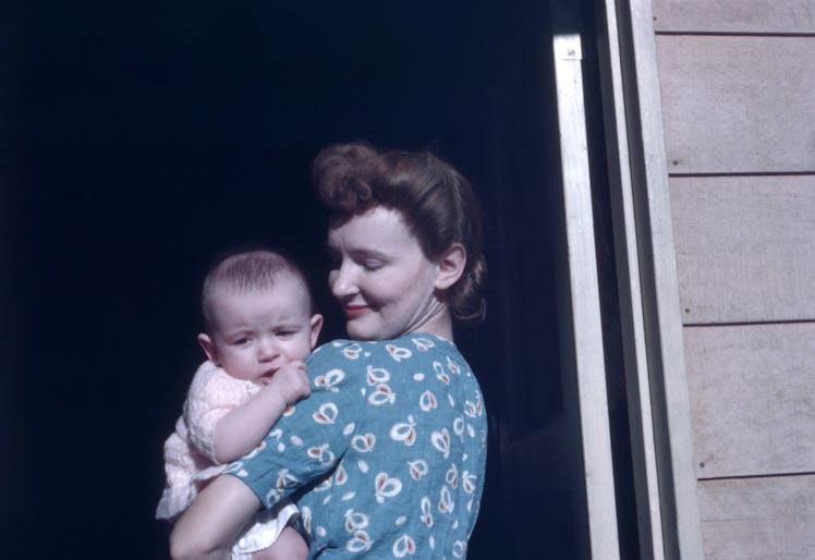 A 35mm film photo shows a mother standing in a doorway with a child in her arms. She gazes lovingly ...