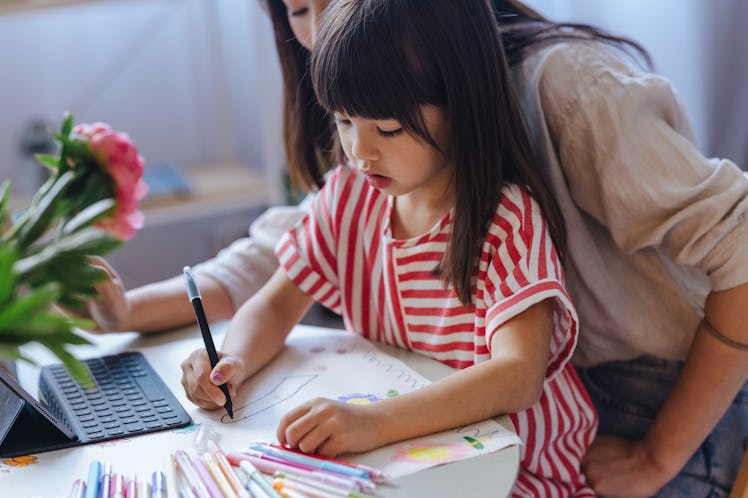 A child sitting in her mother's lap writes on paper, in front of a tablet.