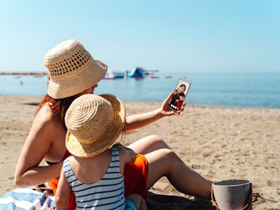 Young Asian mother and daughter using smartphone while having fun on the beach. Summer family vacati...