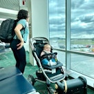 An adorable Eurasian baby boy sits in a stroller and smiles at the camera while his mother and older...
