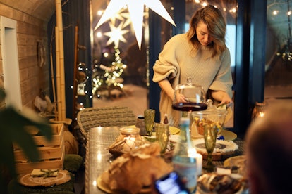 Concentrated young female in warm sweater looking down while standing at dining table with sitting u...