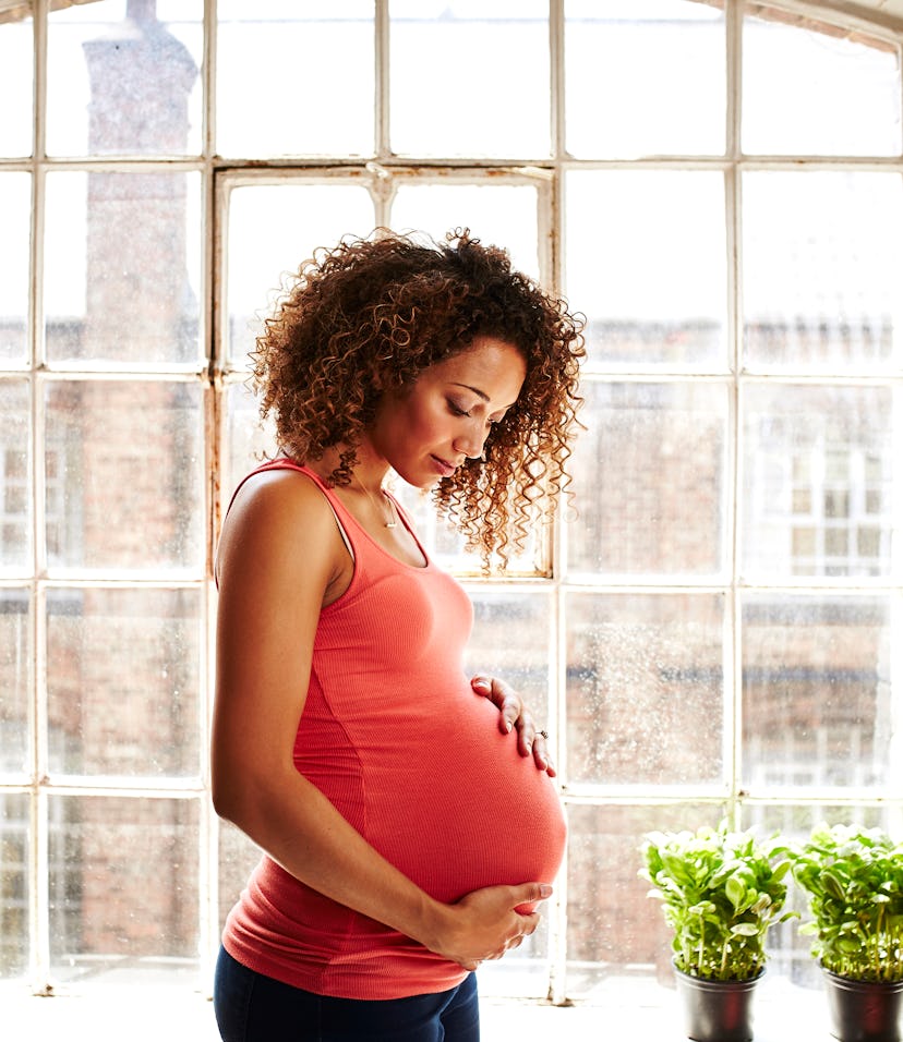 A pregnant woman holds her bump thoughtfully in her modern kitchen window on a sunny day