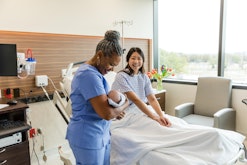 The senior adult female nurse looks down at the newborn in her arms as the mid adult new mother sitt...