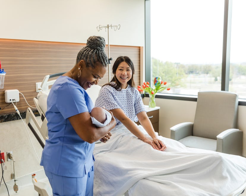 The senior adult female nurse looks down at the newborn in her arms as the mid adult new mother sitt...