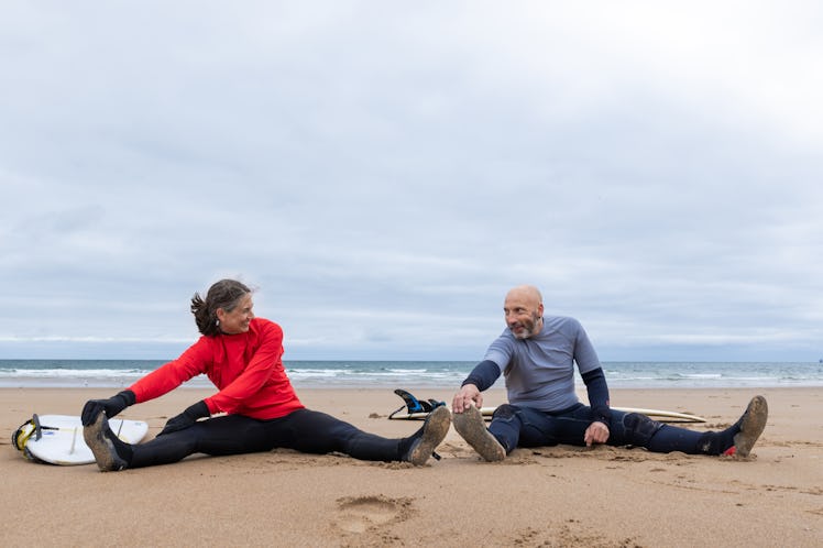A mature couple sitting on Tynemouth Beach and stretching before going surfing. They are both wearin...