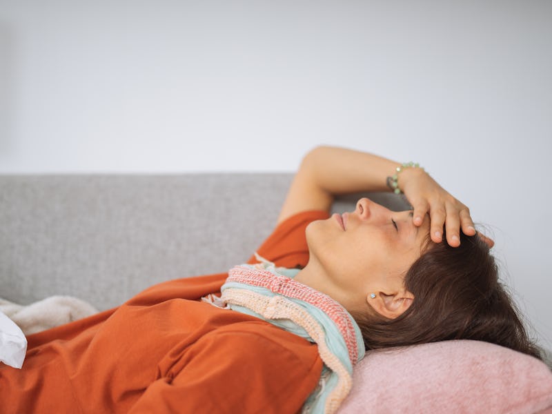 Sick young woman sleeping on living room couch, touching her forehead to see if she has a fever.