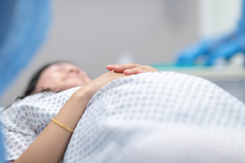 A pregnant woman in a hospital gown lies on an operation table and rests with her hands on her belly...
