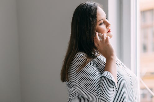 A portrait of a happy overweight woman standing by the window and talking on the phone.