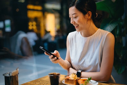 Smiling young Asian woman sitting inside a cozy cafe, scrolling on her smartphone, text messaging wi...