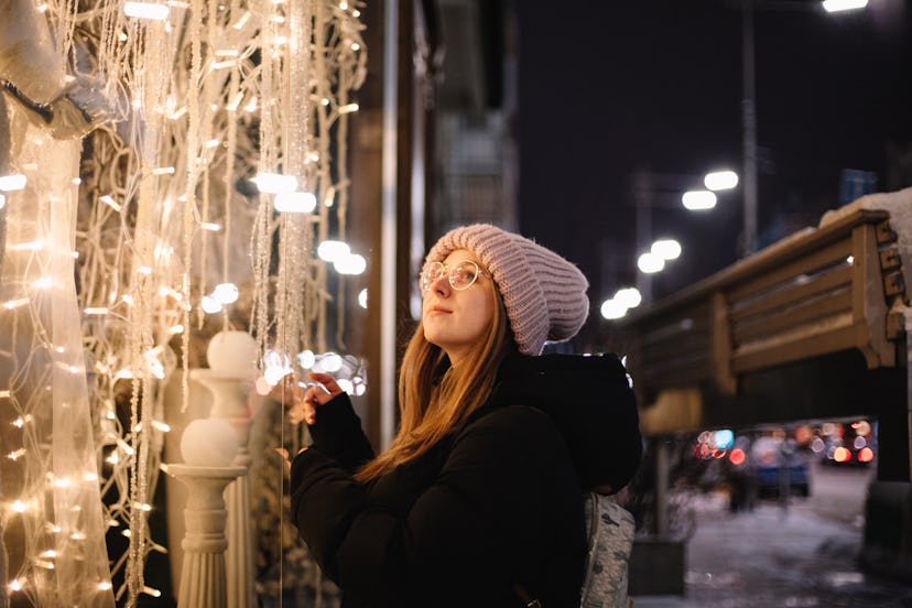 Happy young woman looking at store window decorated with Christmas lights while standing on the stre...