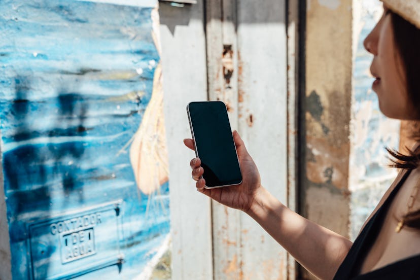 Young woman holding a smartphone with blank screen against a rough wall. Travel and vacation concept...
