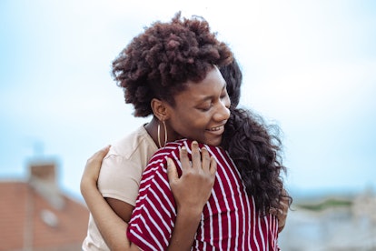 A young African American woman and a young Latin American woman are affectionately hugging on a buil...