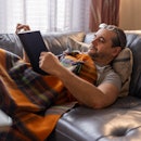 A mature man is resting on a couch, reading a book, covered by a plaid blanket.