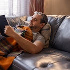 A mature man is resting on a couch, reading a book, covered by a plaid blanket.