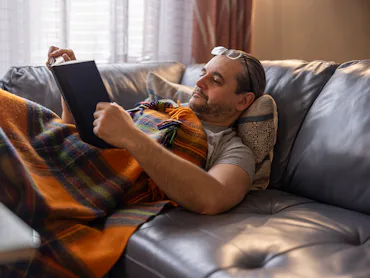 A mature man is resting on a couch, reading a book, covered by a plaid blanket.