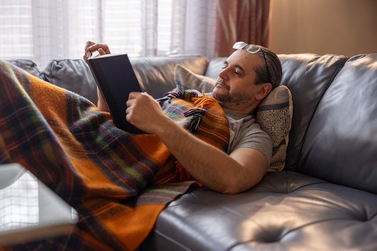 A mature man is resting on a couch, reading a book, covered by a plaid blanket.