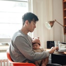 Young Asian father using laptop while holding his baby. Family responsibility. Modern fatherhood.